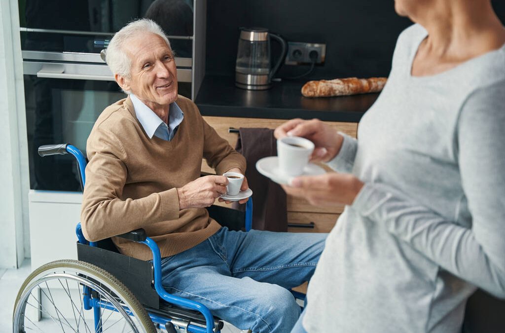 An elderly man in a wheelchair having coffee with a loved one in an accessible kitchen.
